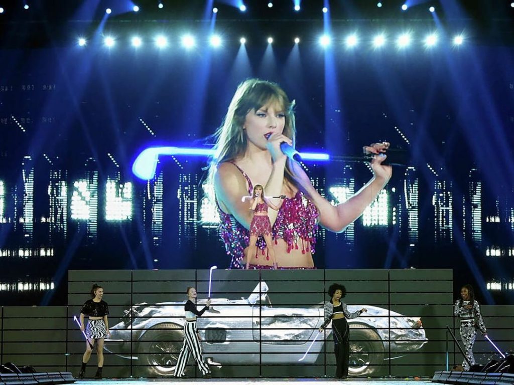 TAYLOR SWIFT SINGING ON STAGE WITH DANCERS BELOW HER AND SILVER CAR ON SCREEN DURING ERAS TOUR